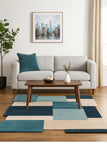 A modern patchwork area rug in shades of teal, navy, and beige is styled in a living room setting on a wooden floor, positioned under a wooden coffee table and a light gray sofa.