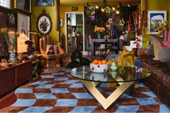 Living room with a brown and blue checkered rug, glass coffee table, hand sculpture chair, and vibrant decor
