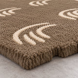 Close-up of a shag area rug in taupe brown with cream-colored abstract wave-like patterns, shown on a light gray concrete floor.