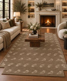 A large rectangular area rug in taupe-brown with a repeating cream wave-like pattern, shown in a styled living room with a sofa, coffee table, fireplace, and wood floor.