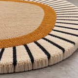 Close-up of a round area rug with a cream center, a mustard-gold ring, and alternating cream and black radial stripes around the edge, shown on a gray floor.