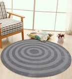 Round grey handwoven wool rug with concentric circles on wooden floor near plaid armchair and floral cushion