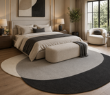 A modern bedroom scene featuring a large oval area rug with a three-tone abstract color-block design in cream, light gray, and charcoal. The rug is styled under a bed with neutral bedding and a matching upholstered bench, with a contemporary armchair and large window in the background.