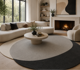 A modern living room featuring a large round area rug with three color-blocked sections in cream, light gray, and charcoal. The rug is styled with a low round stone coffee table, a curved cream sectional sofa, a matching accent chair, and a fireplace in the background.