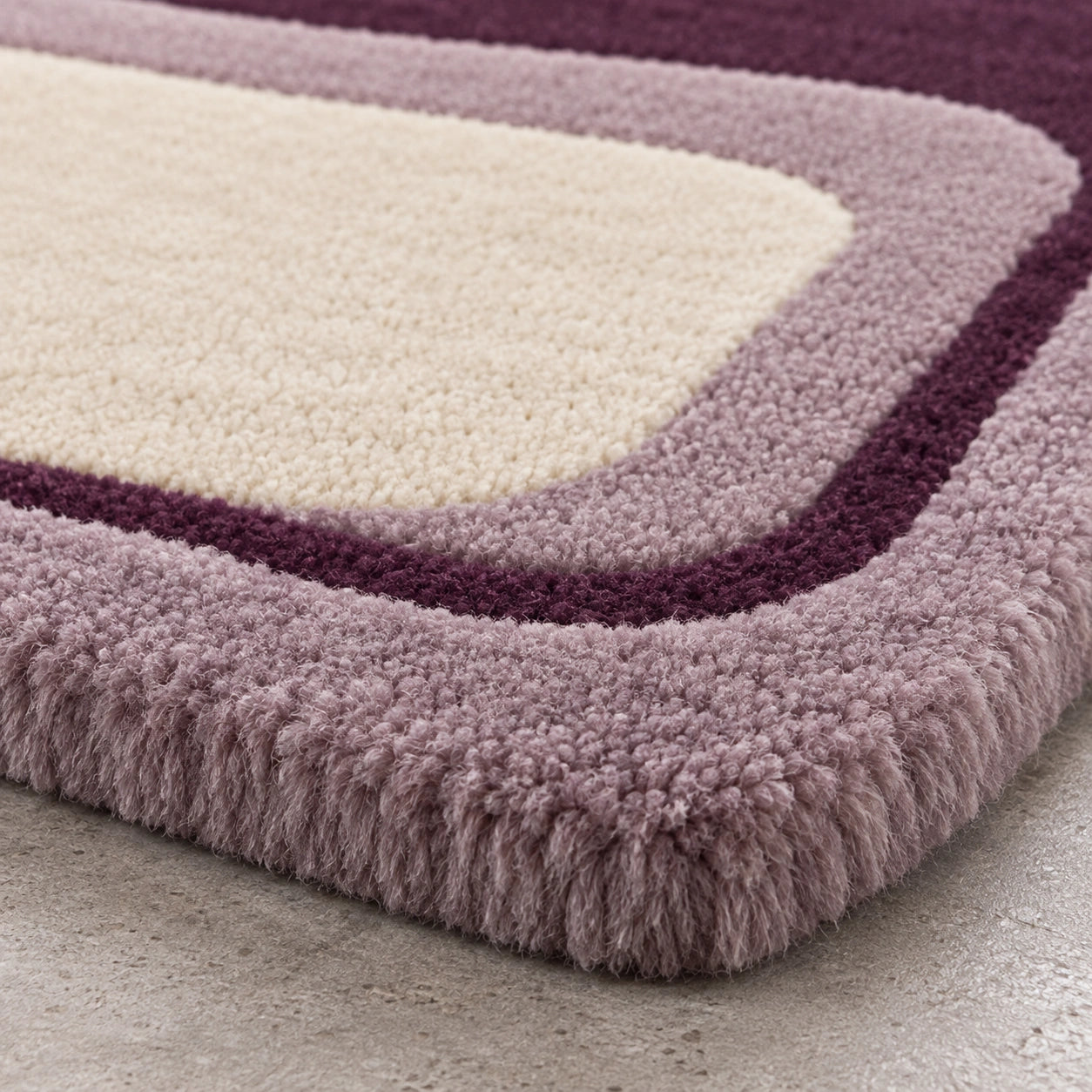 Close-up of a plush shag rug corner showing layered color bands: a cream center, a mauve-purple border, and a dusty lavender edge, photographed on a light concrete floor.