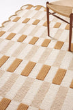 Brown and cream checkered handwoven rug with textured rectangular patterns and scalloped edge under wooden chair