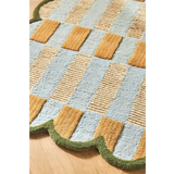 Close-up of a luxury handwoven rug with scalloped green edge, featuring blue, gold, and textured checkered pattern on wooden floor