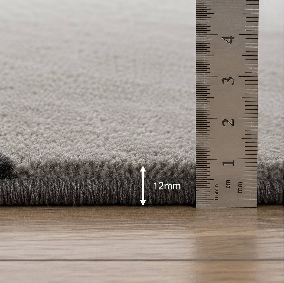 Close-up of a gray textured rug corner with a metal ruler showing a 12 mm pile height, placed on a light wood floor.