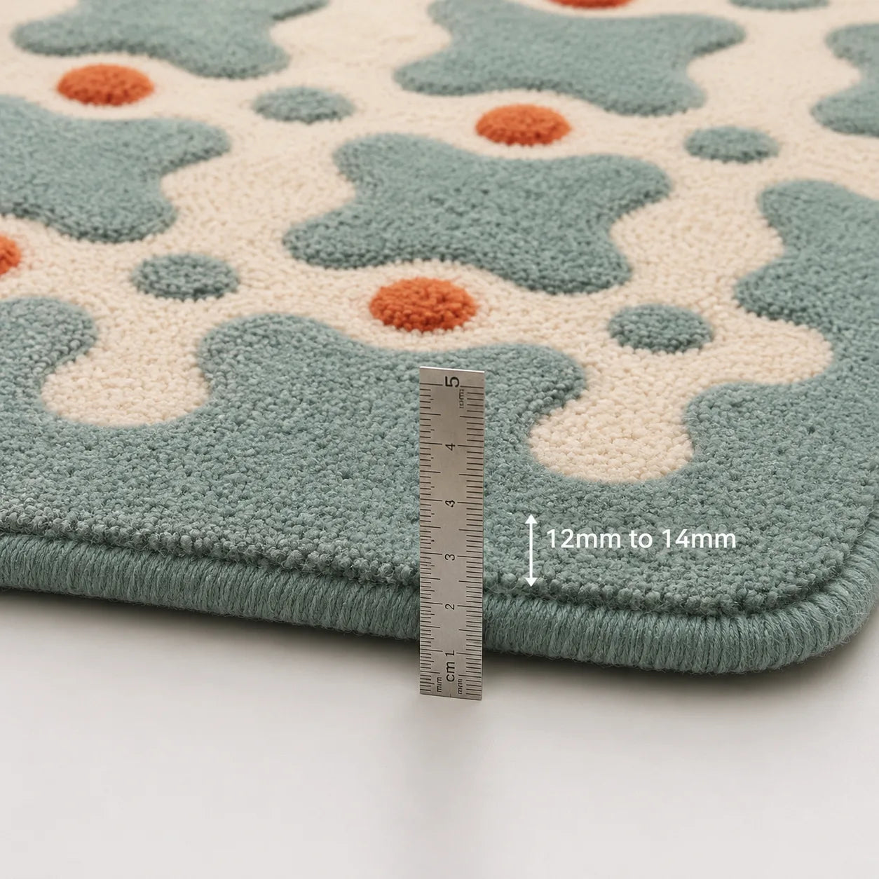 Close-up of a textured rug with a repeating abstract pattern in muted teal and cream, accented with small orange dots. A metal ruler is placed vertically to show the rug's thickness, with text indicating 12mm to 14mm.