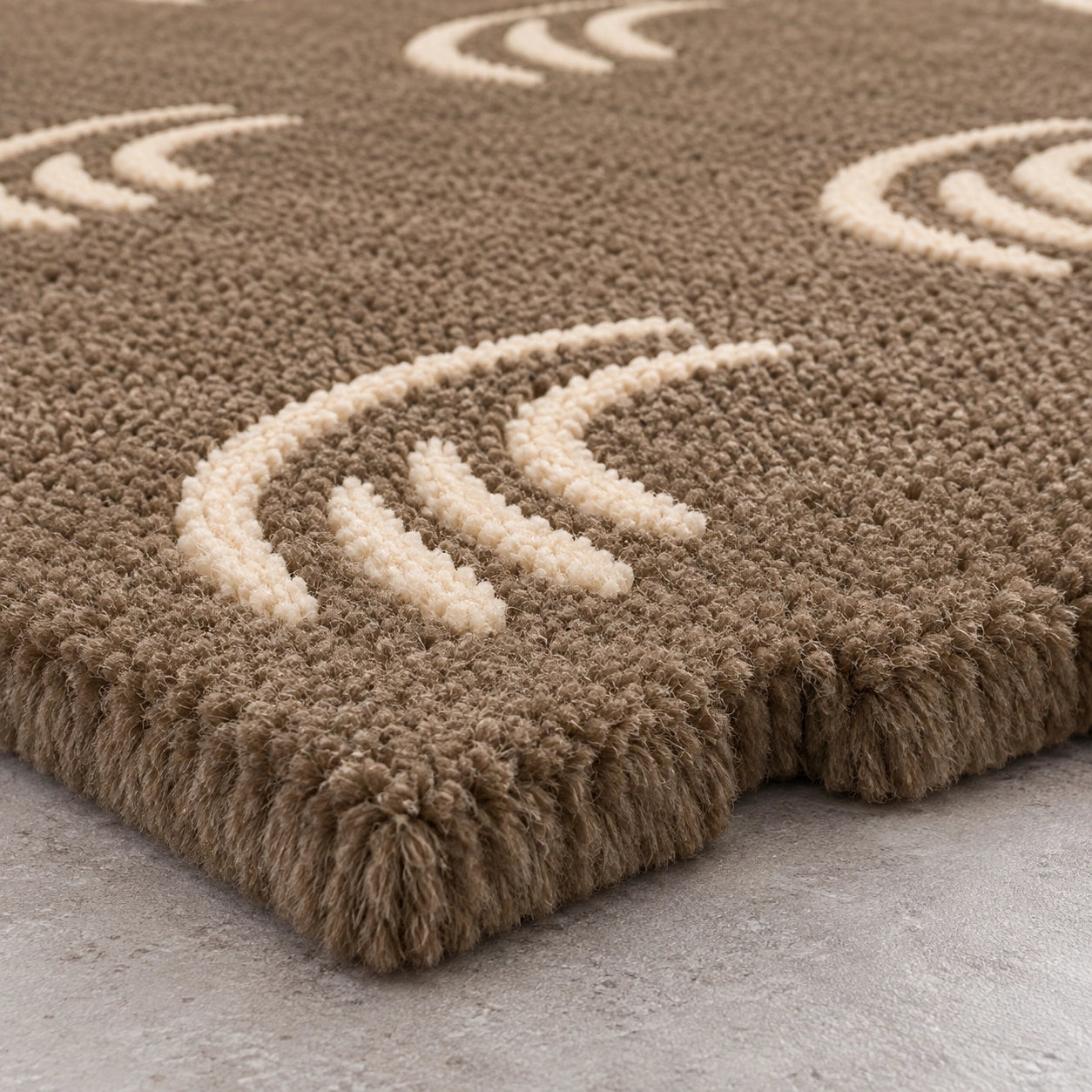 Close-up of a shag area rug in taupe brown with cream-colored abstract wave-like patterns, shown on a light gray concrete floor.
