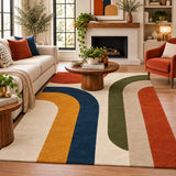 A large area rug with a retro multi-color abstract stripe pattern in navy, mustard yellow, green, rust orange, and cream, styled in a bright, modern living room.
