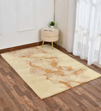 Gold and beige abstract area rug on wooden floor beside beige round ottoman with a plant and book