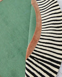 Round handwoven rug with green center, rust border, and black-and-white striped outer edge on marble floor