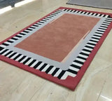 Modern handwoven color block rug with terracotta center, gray inner border, and black-white geometric pattern on red outer edge on marble floor