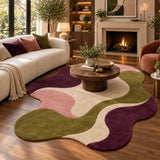 A modern wavy area rug in olive green, cream, pink, and deep purple, styled in a cozy living room with a wooden coffee table, cream sofa, and fireplace in the background.