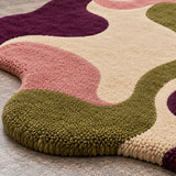 A close-up of a plush, high-pile area rug with a wavy, organic abstract pattern in olive green, dusty pink, cream, and deep purple, shown on a gray stone floor.