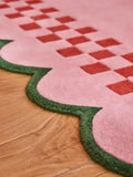 Close-up of pink checkerboard rug with scalloped green edge on wooden floor, luxury handwoven design