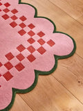 Pink checkerboard rug with scalloped green edge on wooden floor, luxury handwoven design