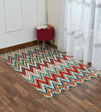 Colorful pixelated chevron rug on wooden floor by red stool with book and cup near white wall and sheer curtains