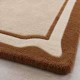 Close-up of a plush area rug corner showing a cream center field with a wavy brown border, photographed on a light neutral floor.