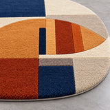 A close-up of a round shag area rug with a modern geometric color-block pattern in navy blue, rust orange, beige, and cream tones, shown on a gray floor.