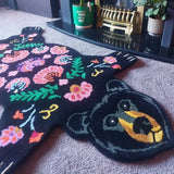 Black bear-shaped rug with colorful floral patterns on beige carpet near fireplace and potted plants