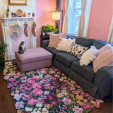 Cozy living room with floral luxury rug, gray sofa with pink and white cushions, pink ottoman with cat, and festive stockings on white mantel