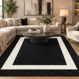 A large rectangular area rug with a black center and wide cream borders, shown in a styled living room with a sofa, coffee table, lamp, and plants.