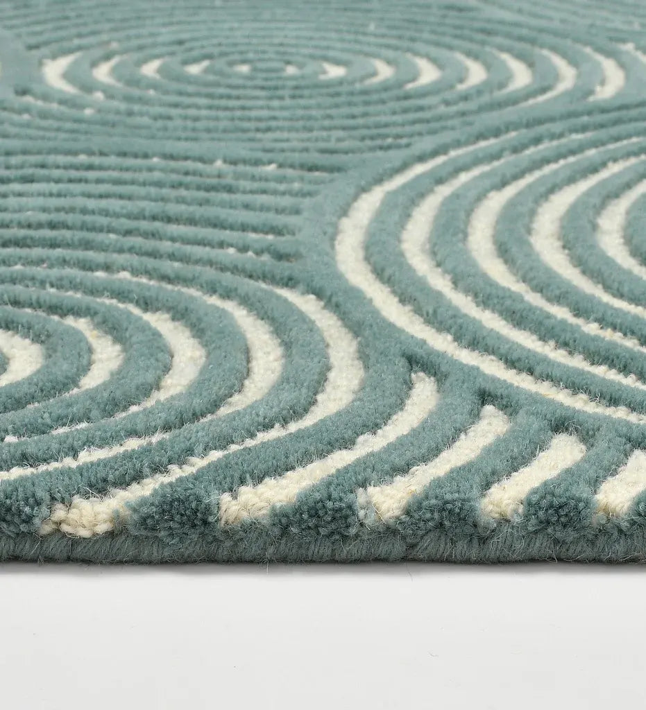 A close-up of a textured area rug with a wavy, concentric spiral pattern in dusty teal and cream, showing the pile texture and the edge of the rug.