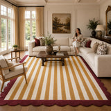 A large striped area rug in mustard yellow and cream with a deep burgundy scalloped border, shown in a bright, modern living room with a woman sitting on a cream sectional sofa.
