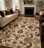 An indoor living room scene showing a large rectangular area rug with an ornate floral and scroll pattern in cream, red, olive green, and taupe. The rug is placed on a dark wood floor between a cream sofa, a brown leather armchair, and a fireplace.