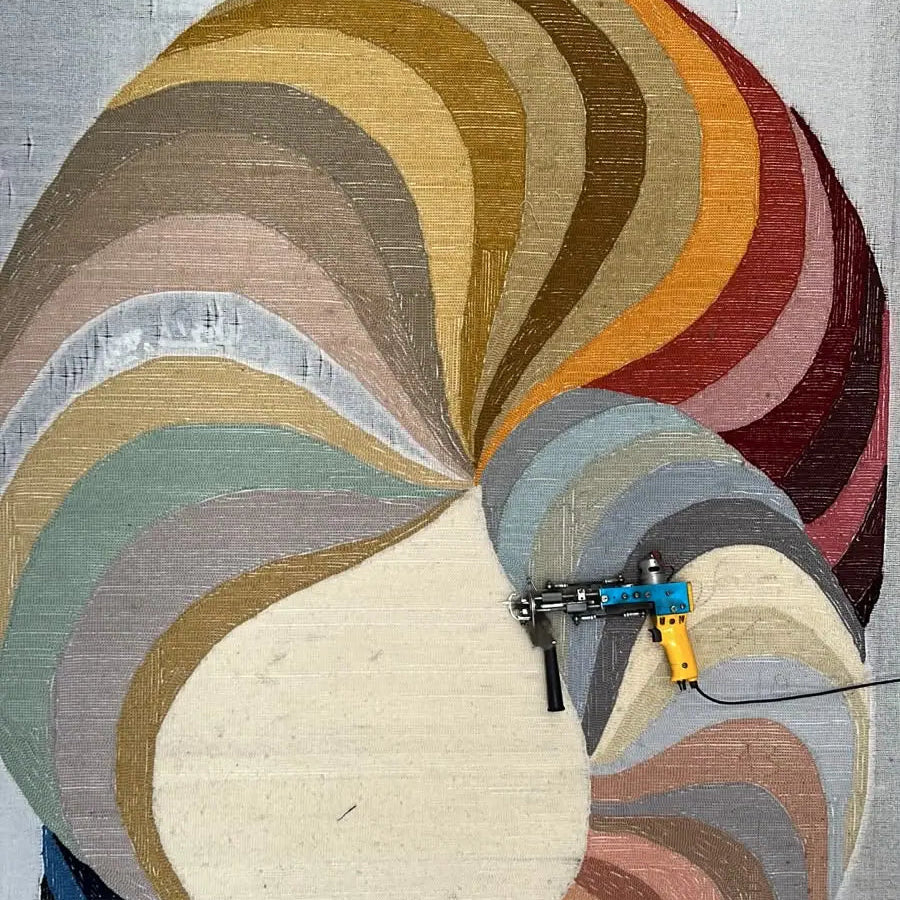 A large abstract textile wall hanging or rug with layered curved bands in cream, beige, brown, gray, blue, orange, red, and pink, shown flat against a light fabric background with a small handheld tool placed near the center.