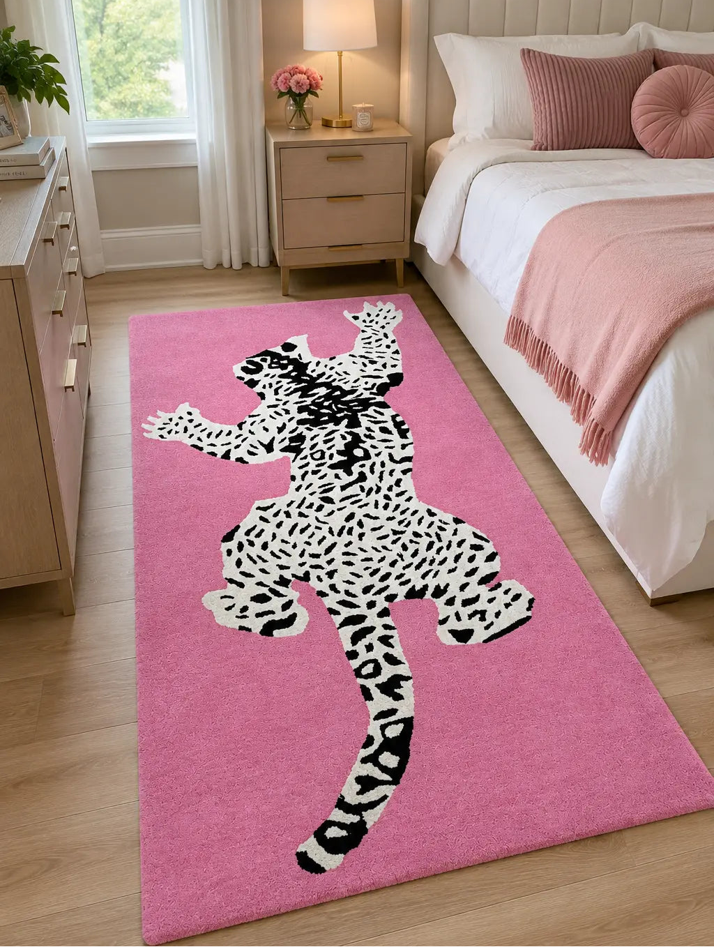 A pink rectangular area rug with a large black-and-white spotted animal silhouette design, shown on a light wood floor in a styled bedroom next to a bed and nightstand.