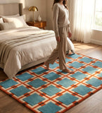 A woman in beige pajamas walks barefoot across a geometric area rug in a bright, modern bedroom. The rug features a pattern of teal blue squares outlined in orange, separated by cream-colored lines, and set on a light wood floor next to a bed with neutral bedding.