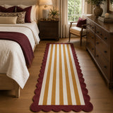 A long runner rug with vertical cream and mustard yellow stripes and a scalloped burgundy border, shown in a styled bedroom setting.